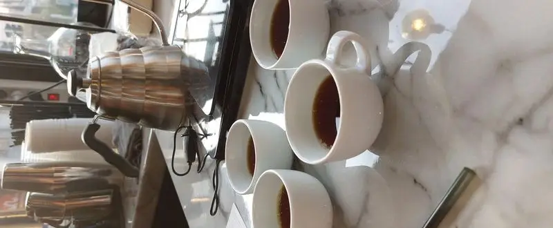 تصویری با کیفیت از فرآیند آماده‌سازی و سرو قهوه دمی خوش‌طعم در کافه آ با کتری استیل گوسنک و فنجان‌های سفید.