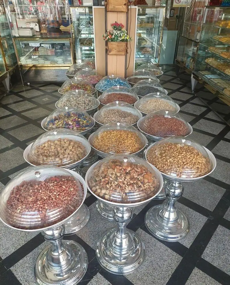 نمایی از شیرینی فروشی باران با انواع آجیل و خشکبار تازه و مرغوب شامل پسته، بادام و بادام زمینی در ظرف‌های نقره‌ای.