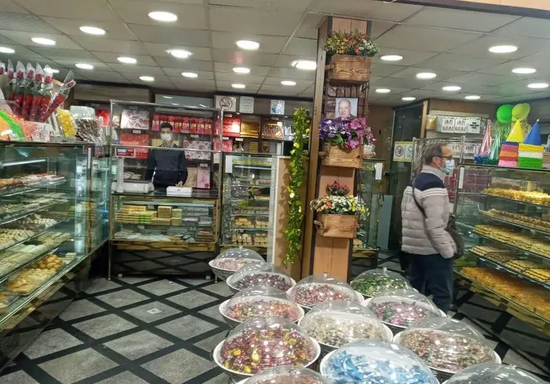 فضای داخلی شیرینی فروشی باران با ویترین‌های بزرگ پر از انواع شیرینی‌های خوشمزه، دسر و آجیل تازه، همراه با کارمند و مشتری ماسک‌دار.