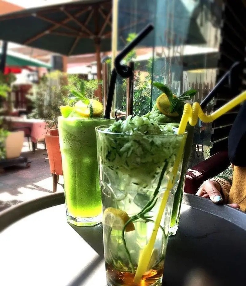 نوشیدنی‌های خنک و تازه کافه لند: اسموتی سبز با لیمو و نعنا، و دتاکس واتر خیار و لیمو در فضای باز کافه.