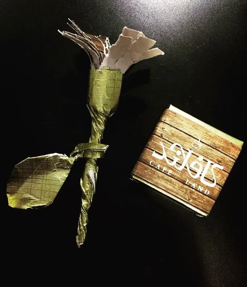 عکس هنری از یک گل دست ساز ساخته شده از فویل طلایی، همراه با بسته شکر برند کافه لند با طرح چوبی زیبا. نمادی از تجربه دلنشین در Cafe Land.