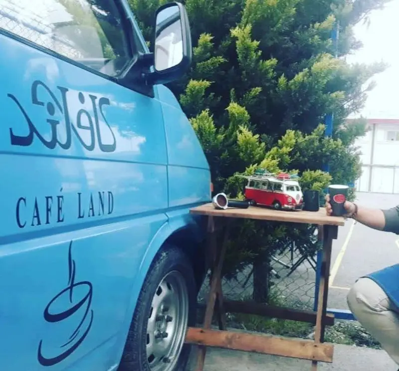 ون آبی کافه لند با نام فارسی و انگلیسی، میز چوبی قهوه با فولکس واگن مینیاتوری. یک دست فنجان قهوه بیرون‌بر در کنار کافه سیار.