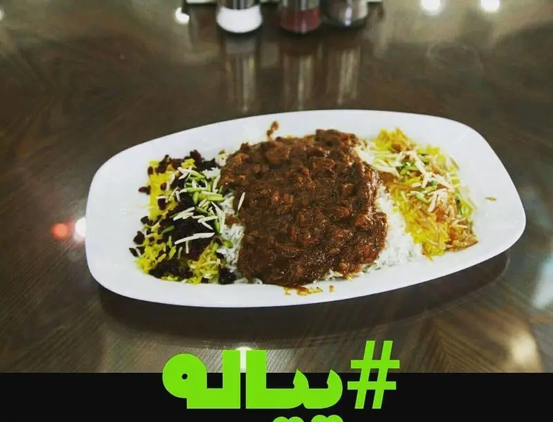 یک بشقاب غذای اصیل ایرانی شامل خورش لذیذ با برنج زعفرانی تزئین شده با زرشک و خلال پسته، آماده سرو در رستوران پیاله.