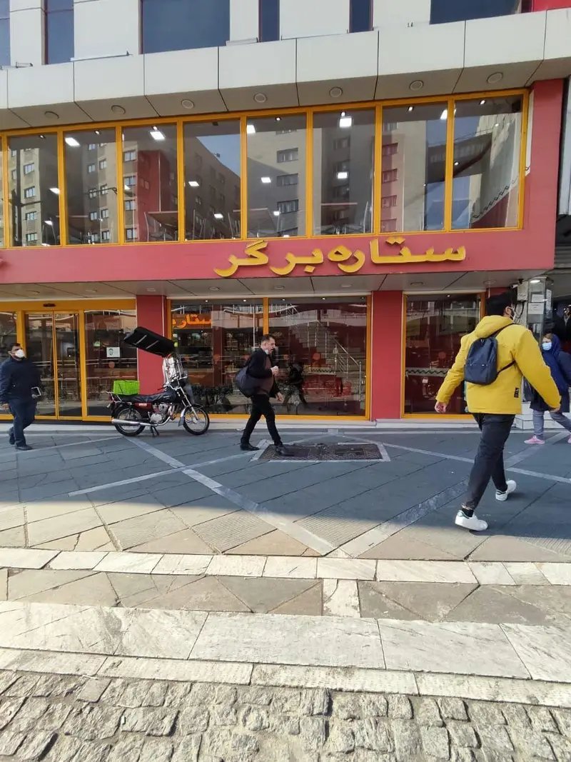 نمای بیرونی رستوران فست فود استار برگر در یک روز آفتابی با حضور مشتریان و یک موتور سیکلت در جلوی درب.