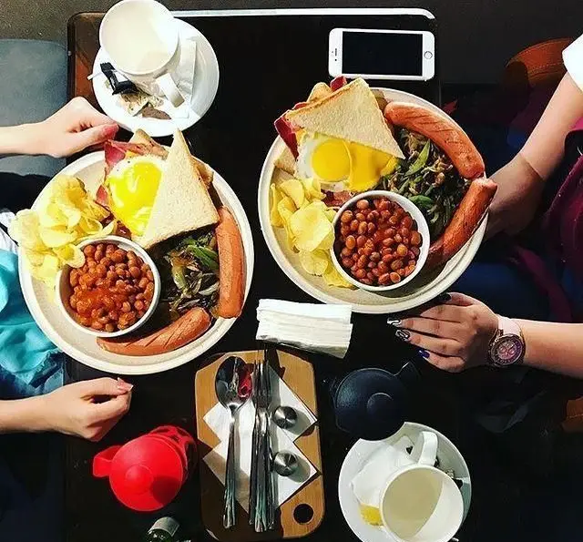 مشتریان در حال لذت بردن از صبحانه انگلیسی کامل و خوشمزه در کافی شاپ تی شن؛ شامل سوسیس، تخم مرغ، لوبیا و نان تست.
