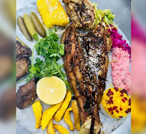 یک پرس کامل و جذاب ماهی کبابی با سس مخصوص، همراه زرشک پلو، سیب زمینی، خیارشور و دورچین سبزیجات تازه در رستوران آروند.