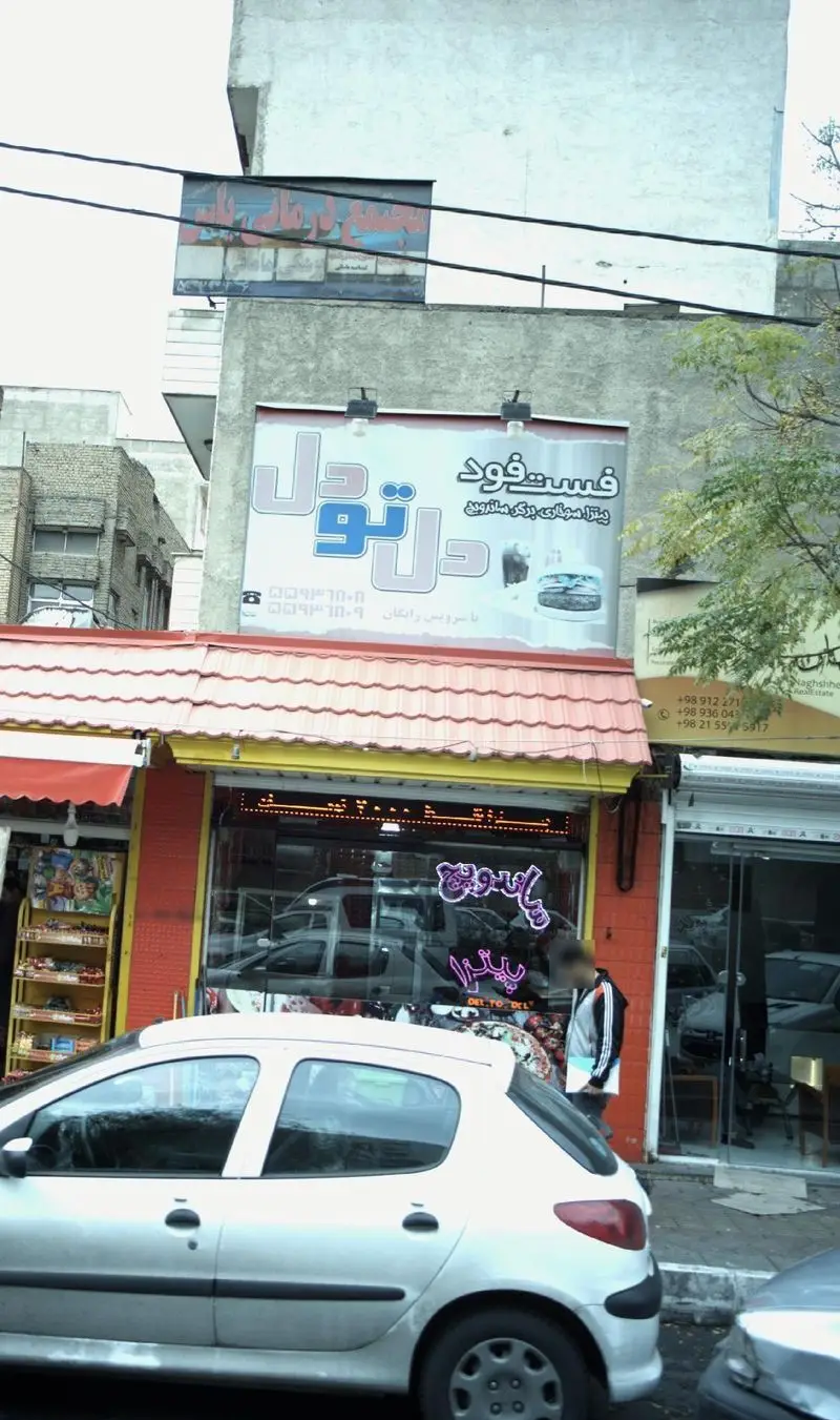 نمای بیرونی فست فود دل تو دل، رستورانی شلوغ در خیابان با تابلوهای پیتزا، ساندویچ و همبرگر. ماشین پژو 206 سفید پارک شده است.