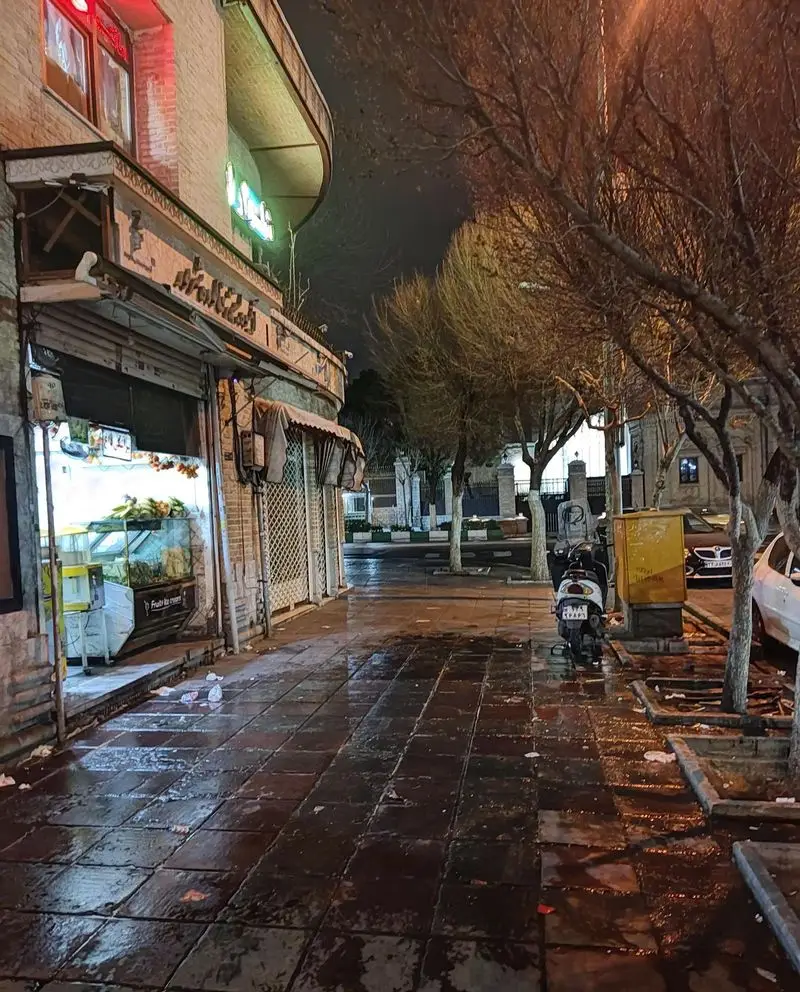 تصویری از آبمیوه‌فروشی شهریور در شب، پیاده‌رو خیس و نورانی، نمای دلنشین خیابان با درختان پاییزی، اسکوتر و ویترین محصولات تازه.