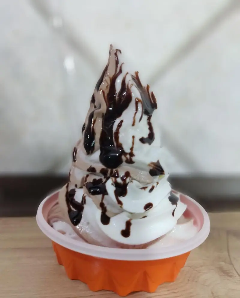 بستنی خوش‌رنگ وانیلی و شکلاتی آبمیوه شهریور با سس شکلات غلیظ، در ظرف نارنجی. ایده‌آل برای روزهای گرم و لحظات شیرین شما.