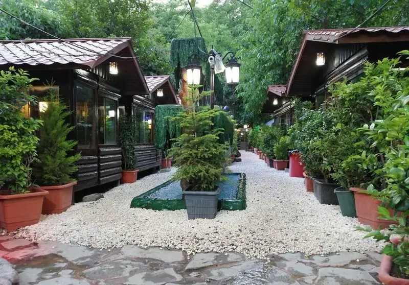 باغ رستوران کلبه: نمای ورودی زیبا با کلبه‌های چوبی دنج، مسیر سنگریزه، گیاهان و درختان سرسبز، فضایی دلنشین برای پذیرایی.