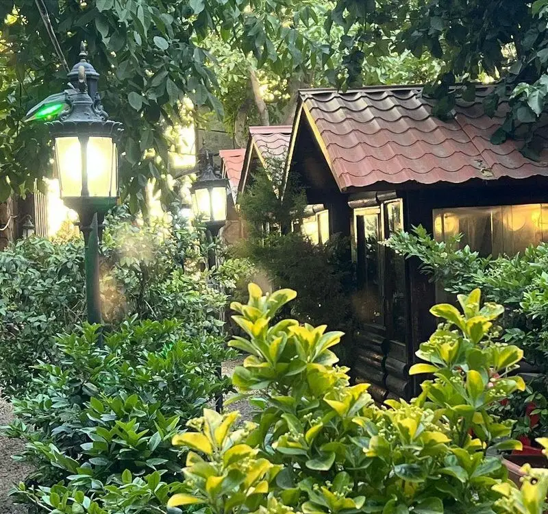 فضای دلنشین باغ رستوران کلبه با کلبه‌های چوبی، درختان سرسبز و نورپردازی زیبای شبانه، محیطی آرامش‌بخش برای مشتریان.