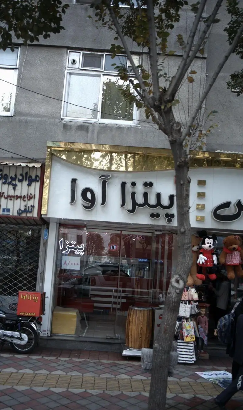 نمای بیرونی جذاب مغازه پیتزا آوا، یک رستوران فست فود شلوغ با تابلوی نئونی، شامل موتور دلیوری و پیاده روی شهری پر جنب و جوش.