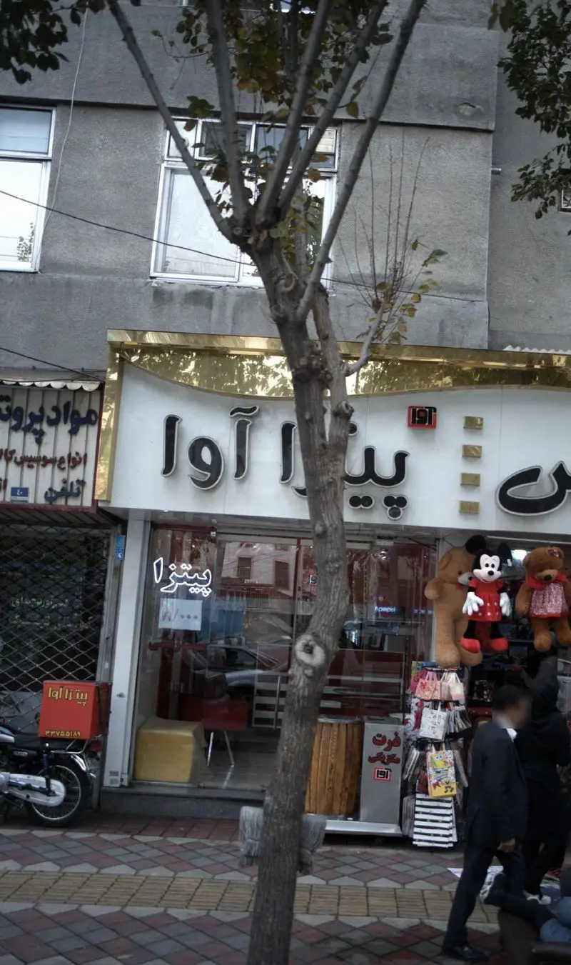 تصویر نمای خیابانی پیتزا آوا، فروشگاه فست فود با تابلوی طلایی بزرگ، موتور پیک دم در و ویترین شیشه‌ای با عروسک‌ها.