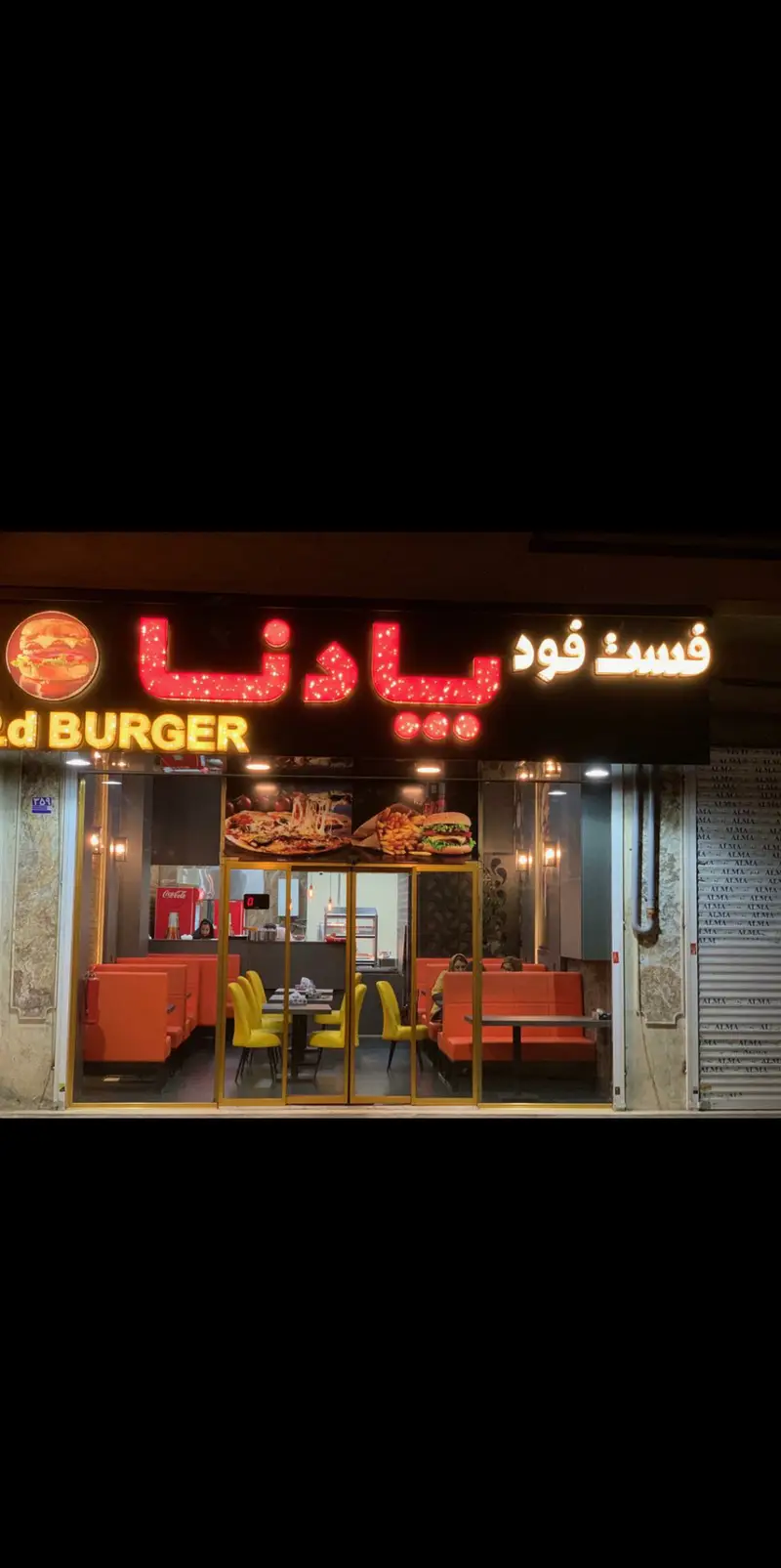 نمای بیرونی فست فود پادنا با تابلوی نئون درخشان، ورودی شیشه‌ای، میز و صندلی‌های نارنجی و زرد و تصاویر جذاب برگر و پیتزا. رستوران مدرن.