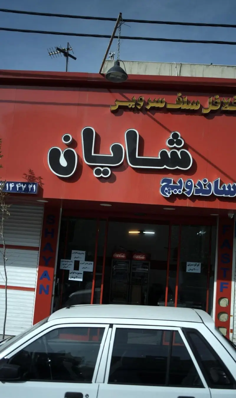 تصویر تابلوی فست فود شایان ارائه دهنده فلافل و ساندویچ سلف سرویس با نمای قرمز رنگ و یک خودرو جلوی ورودی مغازه.