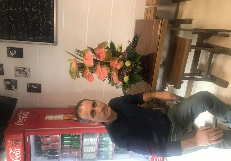 مردی در جگرسرای نیلوفر نشسته است. محیط دلپذیر رستوران با میز، صندلی، گل آرایی، منو و یخچال نوشیدنی مشخص است.