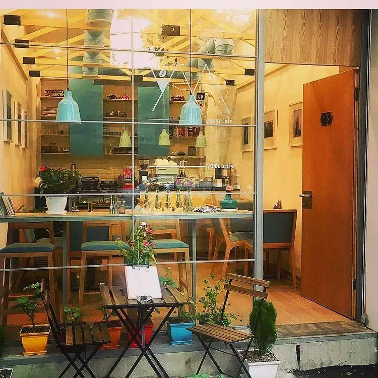 تصویر نمای بیرونی کافه یاما با دکوراسیون مدرن، میز و صندلی‌های فضای باز، فضای دنج داخلی و دستگاه قهوه‌ساز. مکانی ایده‌آل برای نوشیدن قهوه.
