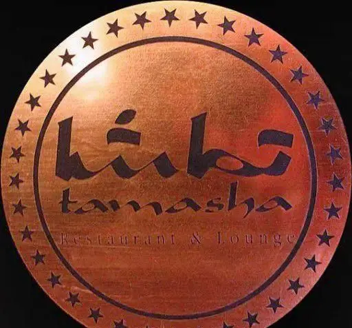 لوگوی مسی و طلایی رستوران تماشا، با خط زیبای فارسی و انگلیسی "Tamasha Restaurant & Lounge" و حاشیه‌ای از ستاره‌های درخشان، نماد برند.