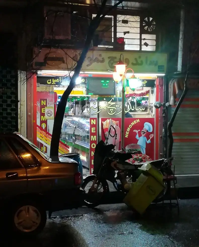 نمای شبانه از فست فود ساندویچ نمونه با تابلوی روشن و لوگوی سرآشپز، همراه با خودرو و موتور سیکلت تحویل غذا در خیابان خیس.