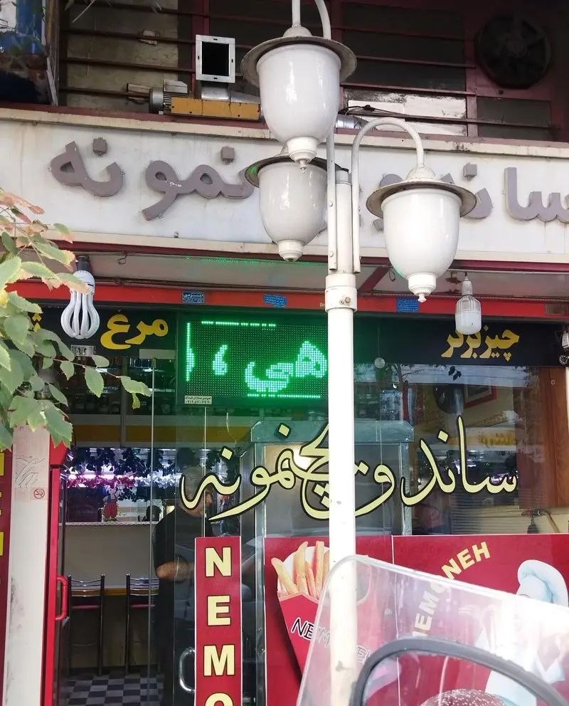 نمای بیرونی رستوران فست فود ساندویچ نمونه با تابلوی اسم مغازه، چراغ‌های خیابانی و تابلوی LED نمایش منو شامل مرغ و چیزبرگر.