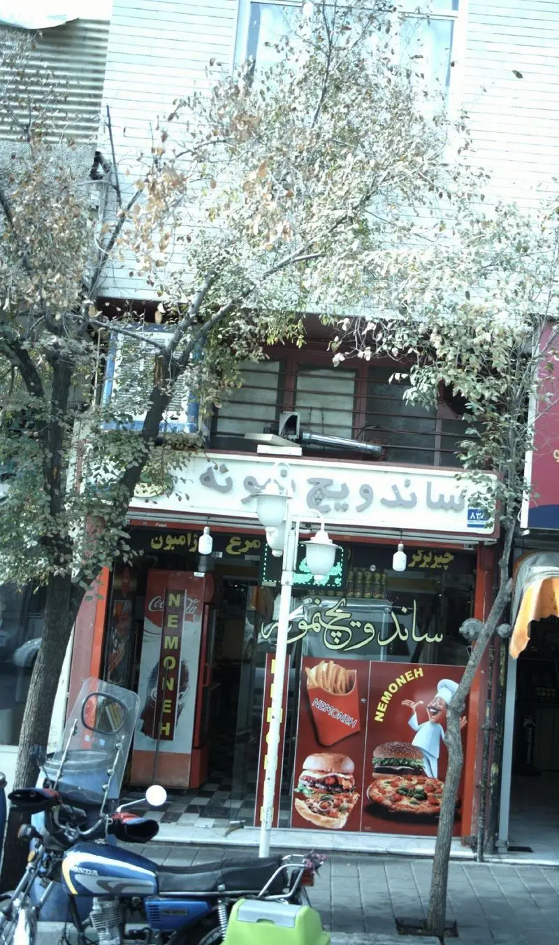 نمای خارجی ساندویچ نمونه، فست فودی با تابلو و ویترین شامل عکس همبرگر، پیتزا و سیب‌زمینی سرخ‌کرده، به همراه موتور سیکلت جلوی مغازه.