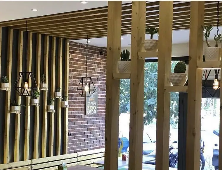 تصویر دکوراسیون داخلی شیک فست فود پاروت شامل دیوار پوش چوبی، آجر نمای قدیمی، لوستر هندسی و پارتیشن چوبی با گلدان‌های طبیعی.