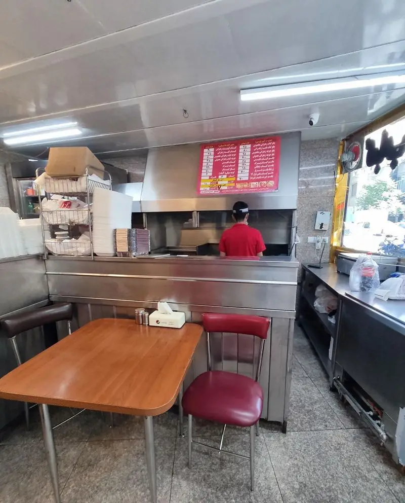 تصویری از فضای داخلی رستوران پاسارگاد شامل پیشخوان آماده سازی غذا، منوی بزرگ بالای سرآشپز، و یک میز ناهارخوری با دو صندلی.