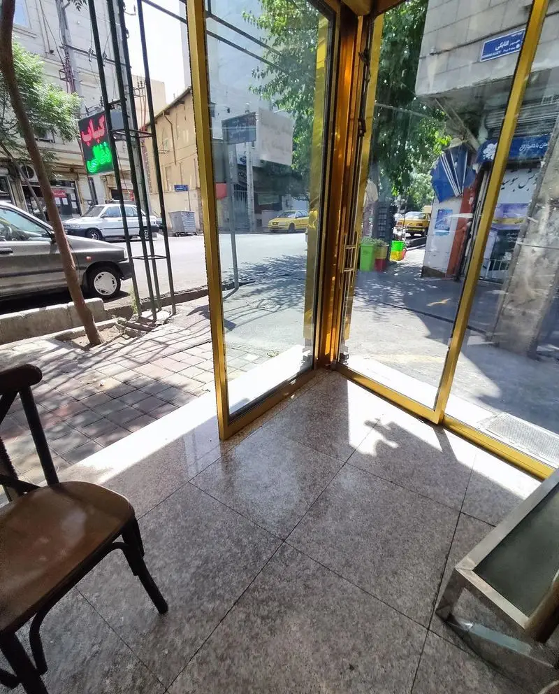 از داخل رستوران پاسارگاد: نمای روشن خیابان با شیشه‌های بزرگ طلایی، تابلوی سبز کباب، خودروها و پیاده‌رو شلوغ.