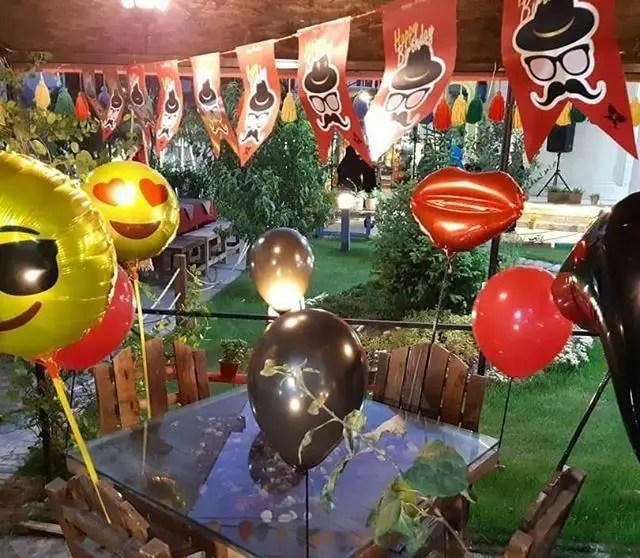 تزیینات زیبای کافه ایل بختیاری برای جشن تولد در فضای باز، شامل بادکنک‌های ایموجی، لب، و سبیل، مکانی دنج برای شب‌های خاص.