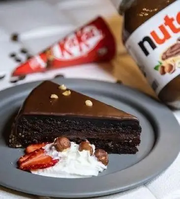 کیک شکلاتی بی‌نظیر کافه نان روتی با روکش براق، فندق، توت فرنگی، خامه. نوتلا و کیت کت در پس‌زمینه.