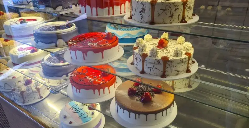 ویترین زیبای قنادی شیرینی سرای پاک پر از انواع کیک‌های تازه و خوشمزه: کیک تولد، شکلاتی، میوه‌ای و دسرهای مجلسی با تزئینات شیک.