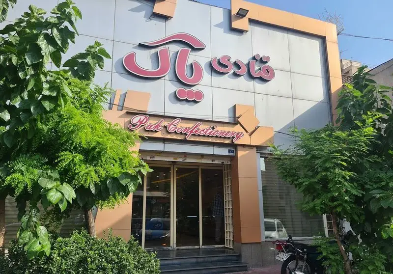 تصویری از نمای بیرونی قنادی پاک (Pak Confectionery) با تابلوهای صورتی و طراحی مدرن خاکستری و نارنجی، و فضای سبز جلوی ورودی.