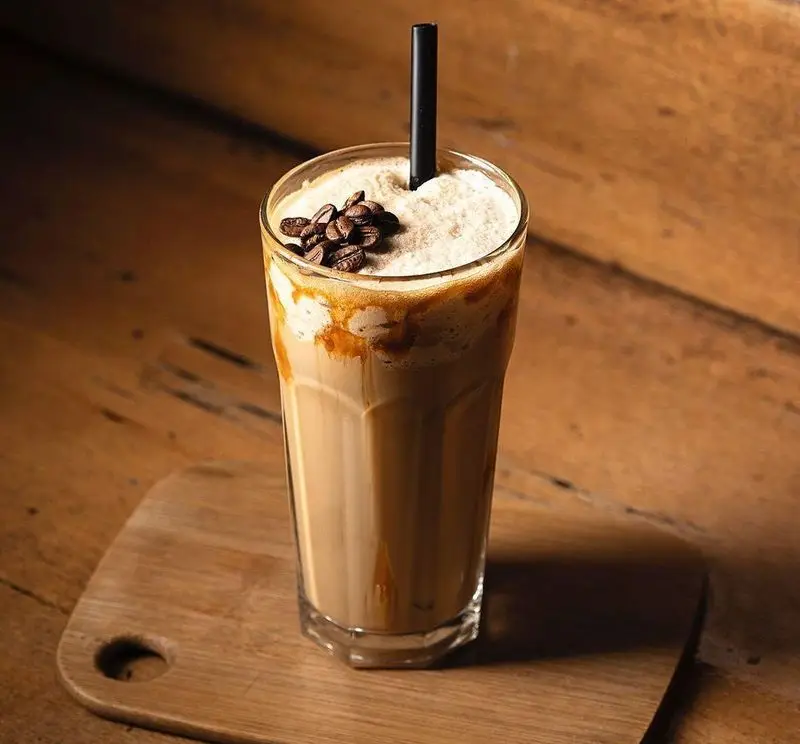 نوشیدنی قهوه سرد کارامل ماکیاتو در لیوان بلند، با فوم و دانه قهوه، از کافه قنادی بیترسوییت.