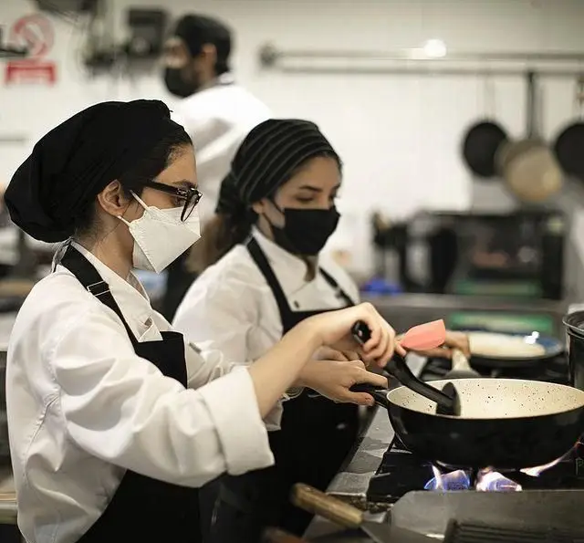 زنان سرآشپز با ماسک و یونیفرم، در حال پخت و پز فعال در آشپزخانه حرفه‌ای کافه قنادی بیترسوییت، رعایت بهداشت و کیفیت.