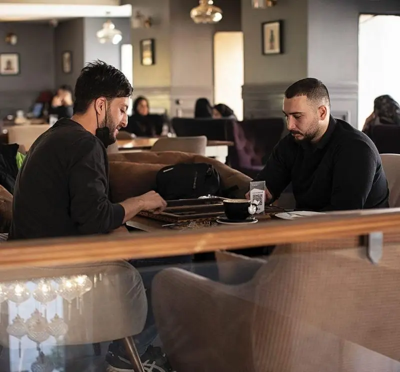 دو مرد در کافه قنادی بیترسوییت مشغول بازی تخته نرد و نوشیدن قهوه، فضایی دوستانه برای اوقات فراغت و دورهمی.