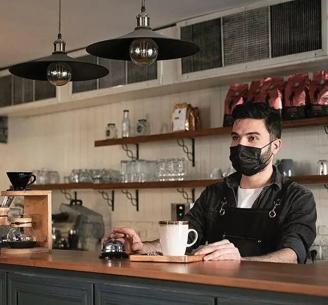 باریستای کافه قنادی بیترسوییت با ماسک مشکی پشت کانتر کافه، آماده سرویس دهی و تهیه انواع قهوه و نوشیدنی های دلپذیر.