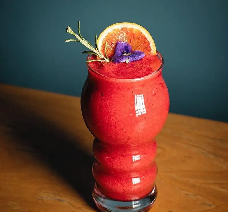 نوشیدنی خوش‌رنگ و لذیذ کافه قنادی بیترسوییت: اسموتی قرمز تازه با تزئین پرتقال خونی، رزماری و گل خوراکی بنفش برای تجربه‌ای خاص.