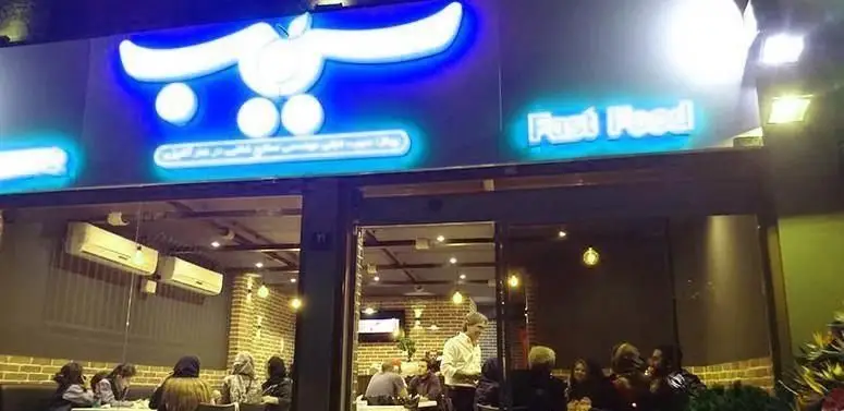 فست فود سیب 360 در شیراز، نمای شبانه با تابلوی آبی و سفید درخشان، ورودی روشن و محیط داخلی دلپذیر با مشتریان، ارائه بهترین فست فود.