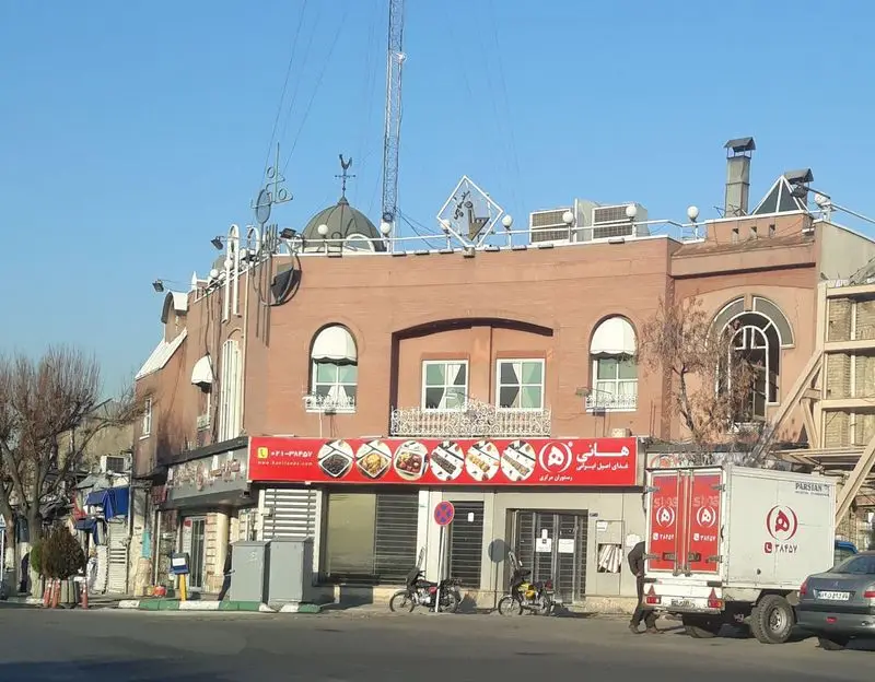 رستوران هانی مرکزی با تابلوی بزرگ غذای اصیل ایرانی و تصاویر غذاها. نمای بیرونی ساختمان در روز روشن، شامل کامیون ارسال و عابران.