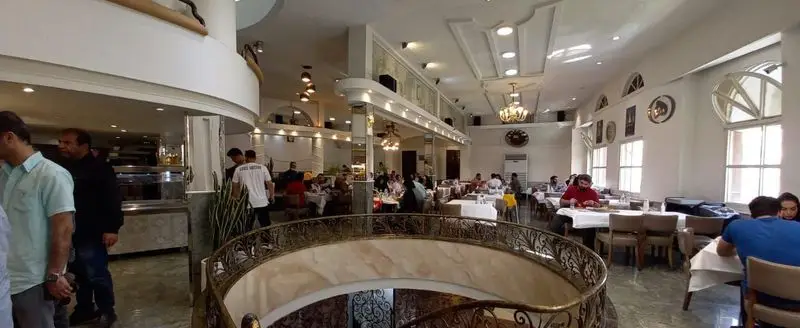 فضای داخلی رستوران هانی مرکزی با دکوراسیون شیک، شامل میز و صندلی‌های پر از مشتریان در حال صرف غذا، و پلکان گردان زیبا.