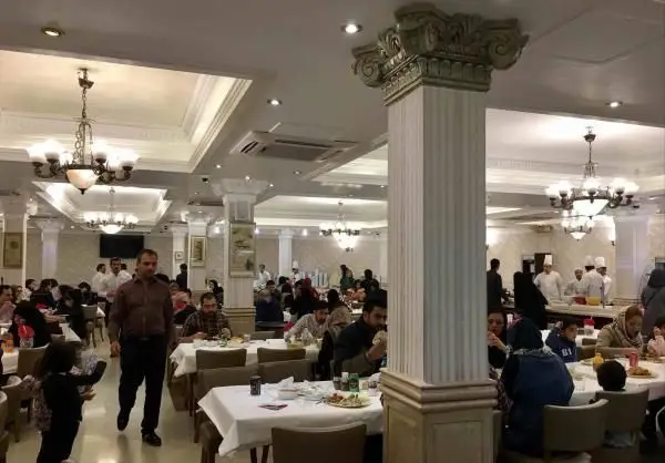 محیط داخلی دلپذیر و پر رفت و آمد رستوران هانی مرکزی با مشتریان مشغول غذا، ستون‌های باشکوه و لوسترهای زیبا.