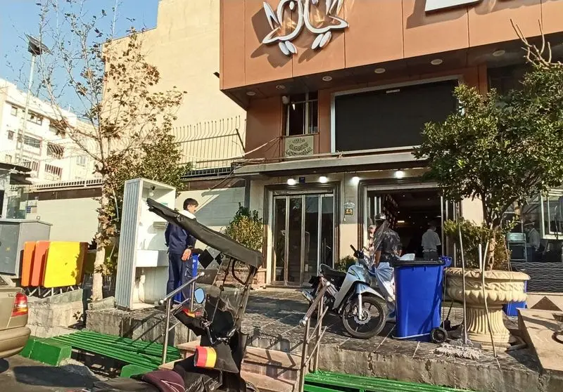 تصویر بیرونی اغذیه نشاط، محلی پرتردد برای تهیه غذا در شهر، با جزئیات خیابان، شامل موتورسیکلت، ماشین و عابران.