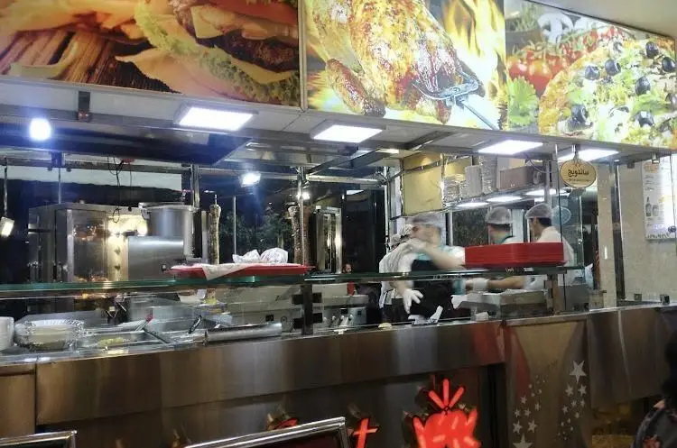 اغذیه نشاط: نمای داخلی آشپزخانه شلوغ و تمیز با پرسنل مشغول تهیه ساندویچ، همبرگر، مرغ بریان و پیتزا. کیفیت بالا و محیطی دلپذیر.