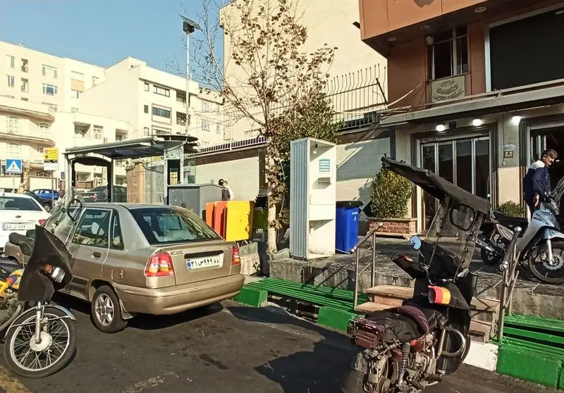 تصویر بیرونی اغذیه نشاط در خیابان‌های تهران، شامل نمای فروشگاه، ماشین‌ها و موتورهای پارک شده در یک روز آفتابی.