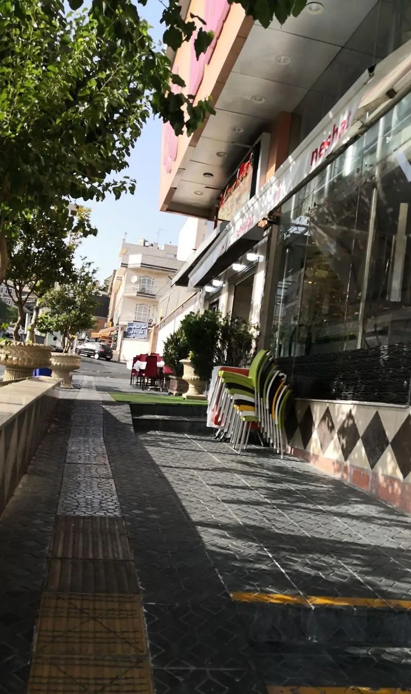 نمایی از ورودی بیرونی اغذیه نشاط در روز روشن، با درختان سرسبز و صندلی‌های رنگی چیده شده آماده برای مشتریان در پیاده‌رو خیابان.