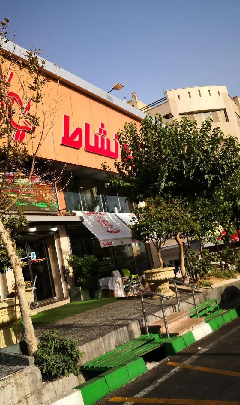 نمای بیرونی و ورودی اصلی اغذیه نشاط با تابلوی بزرگ و قرمز رنگ، محیط دلنشین و سرسبز در کنار خیابان، ایده‌آل برای بازدیدکنندگان.