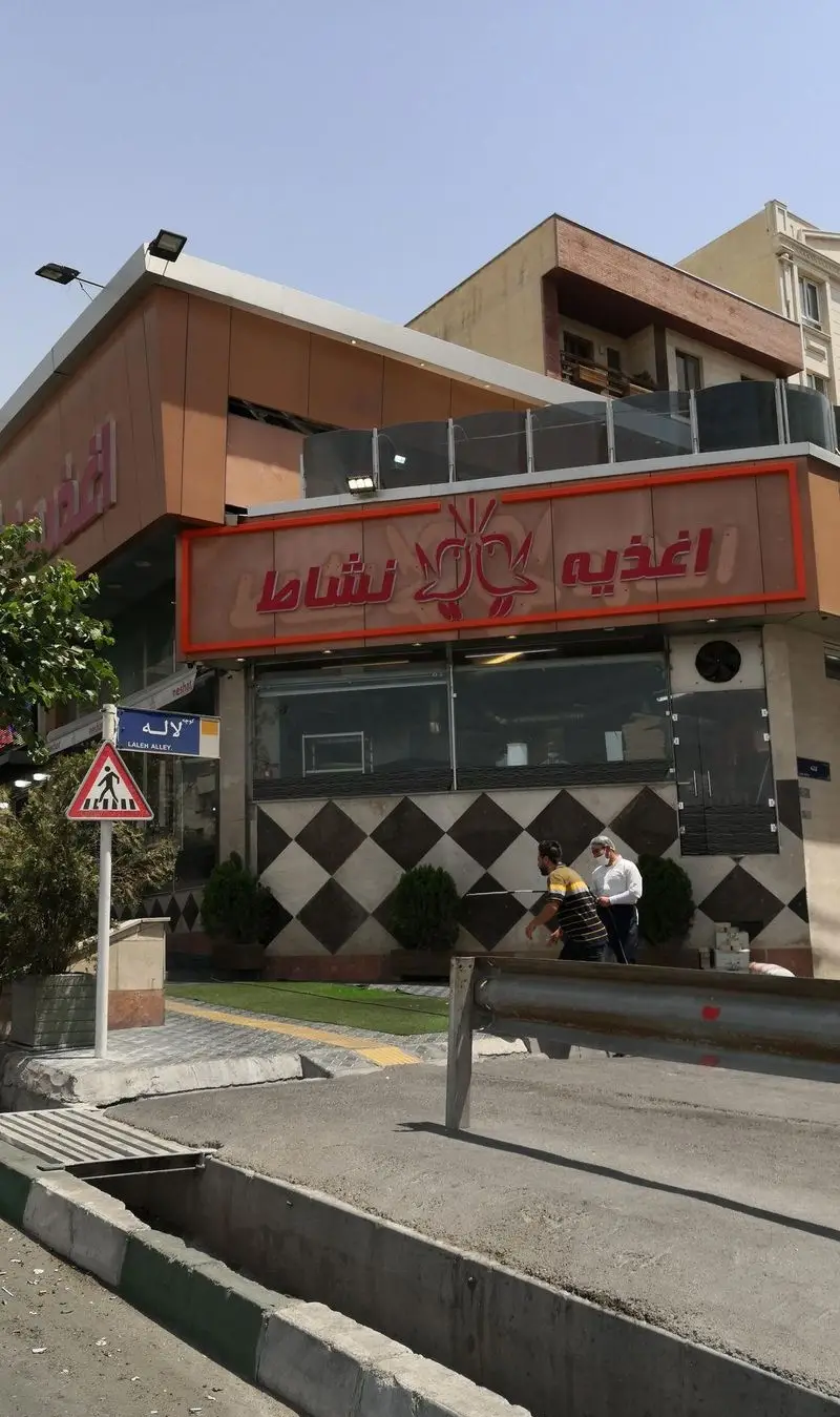 نمای بیرونی اغذیه نشاط در روز روشن، با تابلوی بزرگ و قرمز رنگ، طراحی دیوارهای مربعی و بلوک‌های پیاده‌رو در خیابان لاله تهران.