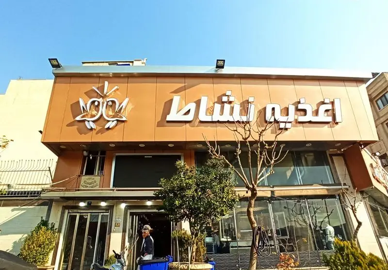 نمای خارجی اغذیه نشاط با تابلوی برجسته نام فروشگاه، لوگوی دو پرنده، ورودی اصلی و محیط خیابان با درختان در کنار ساختمان