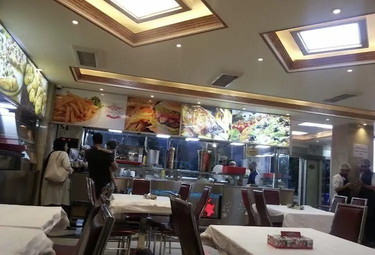 فضای داخلی اغذیه نشاط با میزهای تمیز، مشتریان و پرسنل فعال، منوی غذای متنوع شامل فست فود، ساندویچ و کباب روی تابلوهای بزرگ.