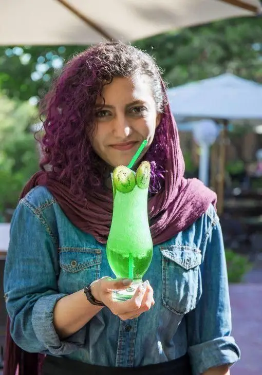 زن جوان با موهای بنفش و شال ارغوانی در باشگاه مینی گلف، نوشیدنی خنک کیوی در دست و لبخند بر لب. تجربه لذت بخش اوقات فراغت.